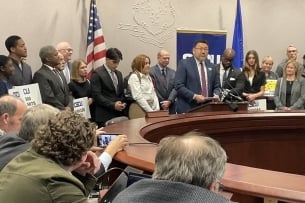 A man in a suit addresses lawmakers backed by supporters